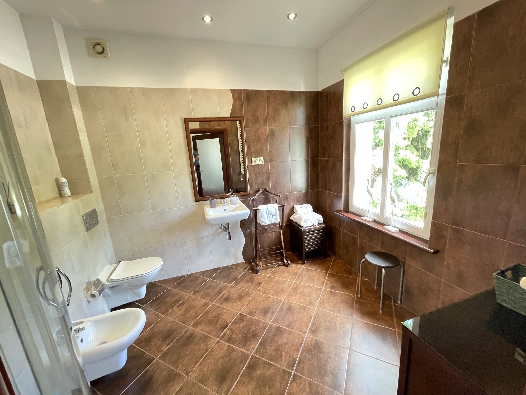 Un baño moderno con inodoro, bidé, lavamanos y ventana con vistas al verdor.