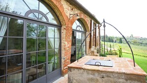 A room with large windows, a stone fireplace, and a view of a vineyard. - Guesthouse 2/4 people, 2 rooms, kitchen, bathroom, patio, A/C, pool, EV charger (San Casciano in Val di Pesa)
