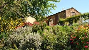 A stone building with arched windows and a brick roof, surrounded by a variety of flowering plants and trees. - Guesthouse 2/4 people, 2 rooms, kitchen, bathroom, patio, A/C, pool, EV charger (San Casciano in Val di Pesa)