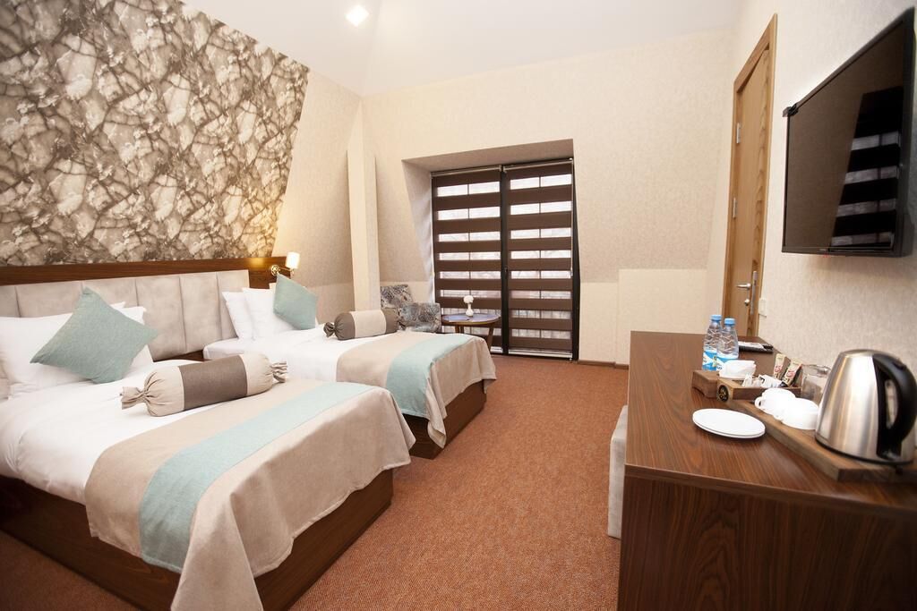 A hotel room with two beds, a wooden desk, a television, and a large window with blinds.