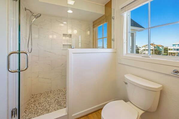 A bathroom with a glass-enclosed shower, a toilet, and a window with a view of houses.