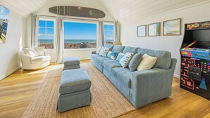 A spacious living room with a large grey sofa, a white armchair, and a small ottoman. A large window offers a view of the beach and ocean. - OCEANFRONT-6 BDRM-4 Mstrs-RENOVATED 2024-Heated Pool,Hot Tub, Media & Game Rooms (Waves)