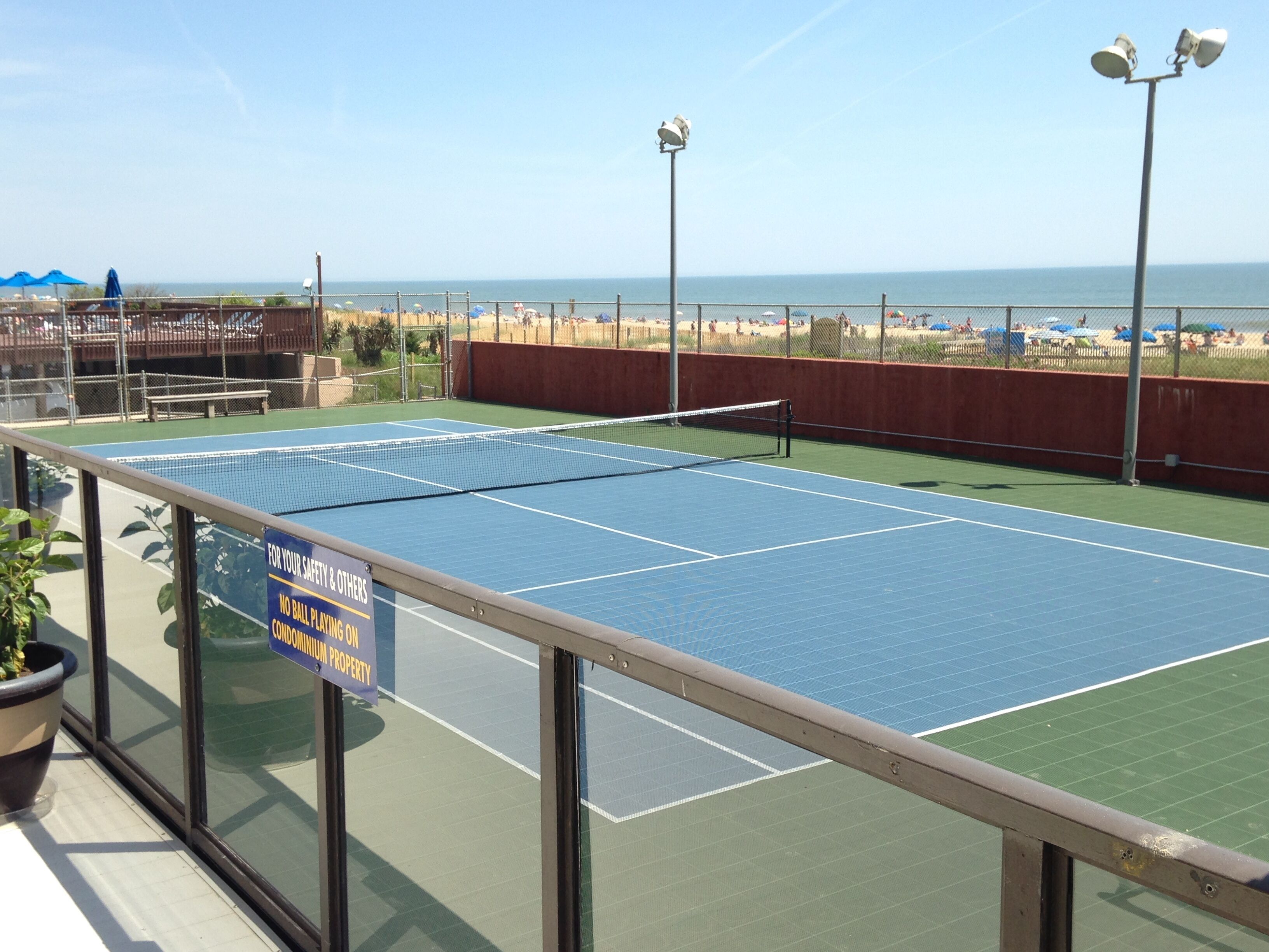 An outdoor tennis court with a safety sign, surrounded by a fence and overlooking a beach.
