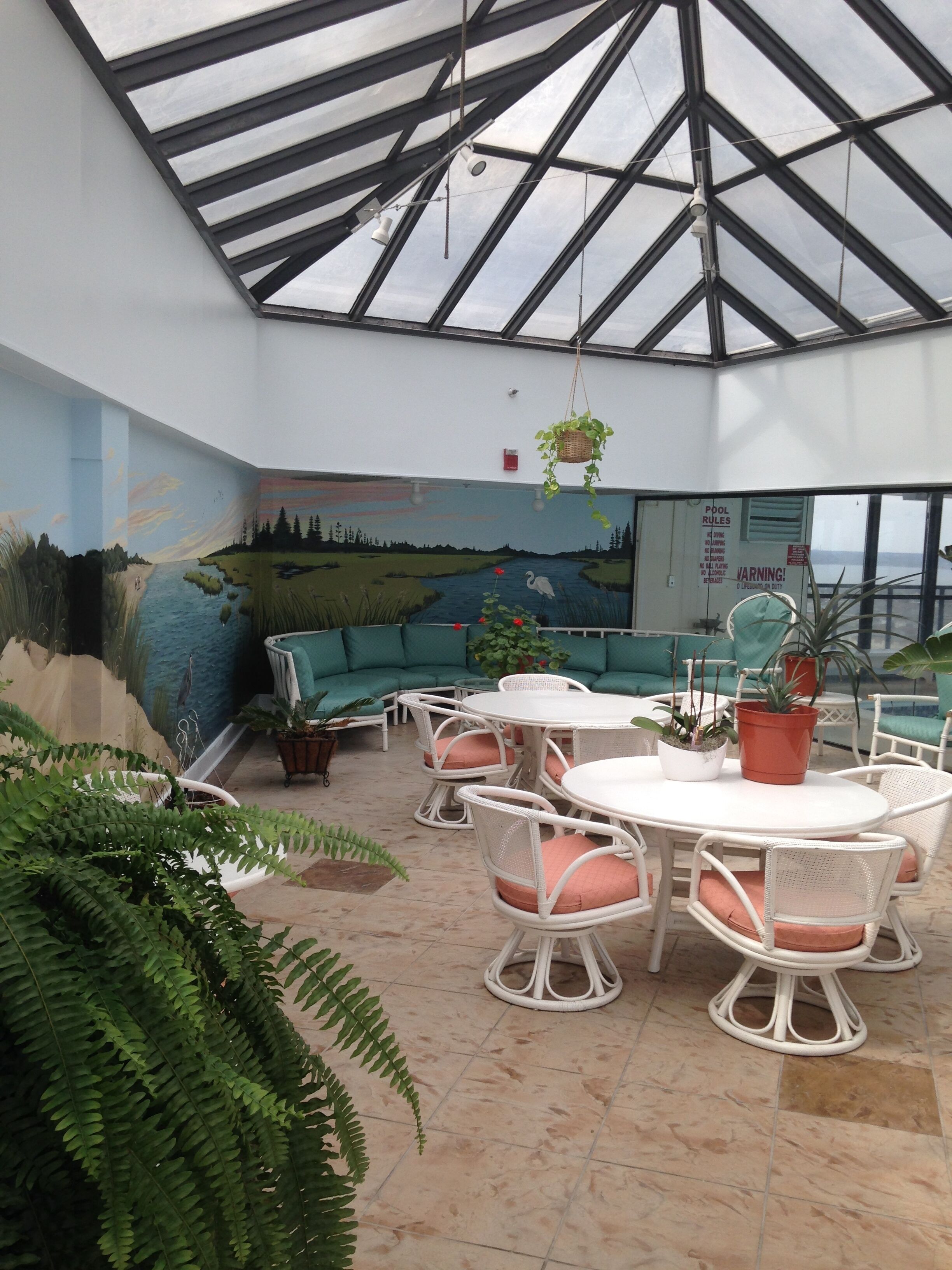 Amplio espacio interior con techo alto, un mural de un paisaje, una piscina con normas y un área de estar con mesas y sillas.