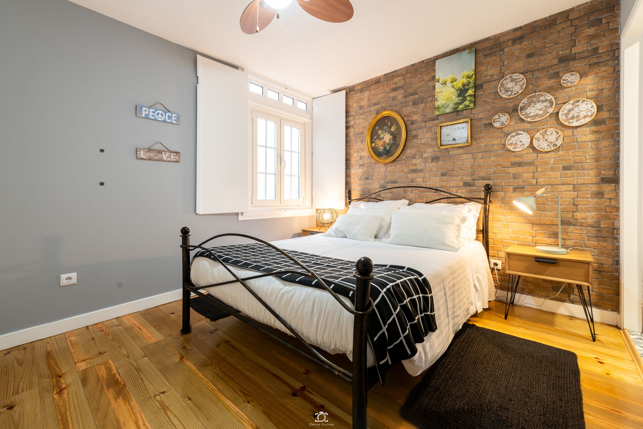 A bedroom with a bed, a nightstand, a lamp, a window, and a brick wall.