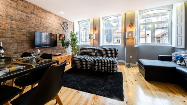A modern living room with a brick wall, a flat-screen TV, a black sofa, a dining table with chairs, and large windows with arched tops. - Merc Porto Downtown (Porto)