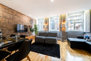 En moderne stue med en murstensvæg, et fladskærms TV, et spiseområde med et bord dækket til to personer, og en sort sofa.