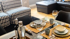 A dining area with a table set for two, including plates, glasses, and cutlery. There are two bottles on the table, and a woven placemat under the plates. The room has a modern design with a black sofa and patterned cushions in the background. - Merc Porto Downtown (Porto)