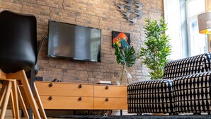 A living room with a brick wall, a flat-screen TV, a wooden cabinet, a black chair, a patterned sofa, and a plant. - Merc Porto Downtown (Porto)