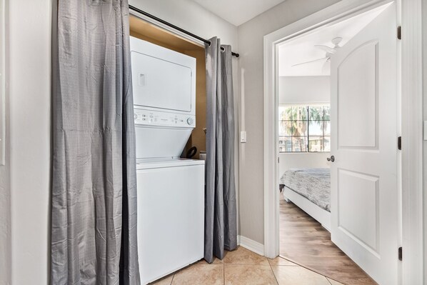 A laundry room with a washing machine, grey curtains, and a bed in the adjacent room.