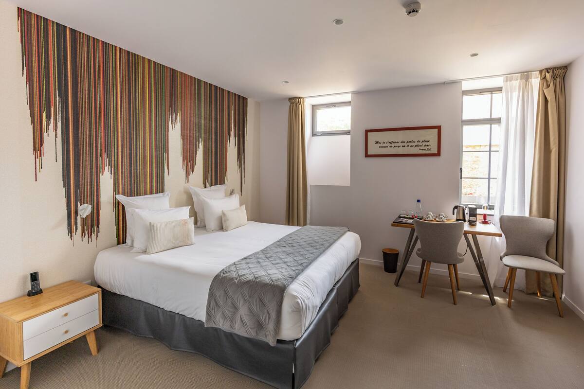 Hotelkamer met een bed, een bureau, twee stoelen, een nachtkastje, een raam met gordijnen en een muur met een kleurrijk abstract ontwerp.