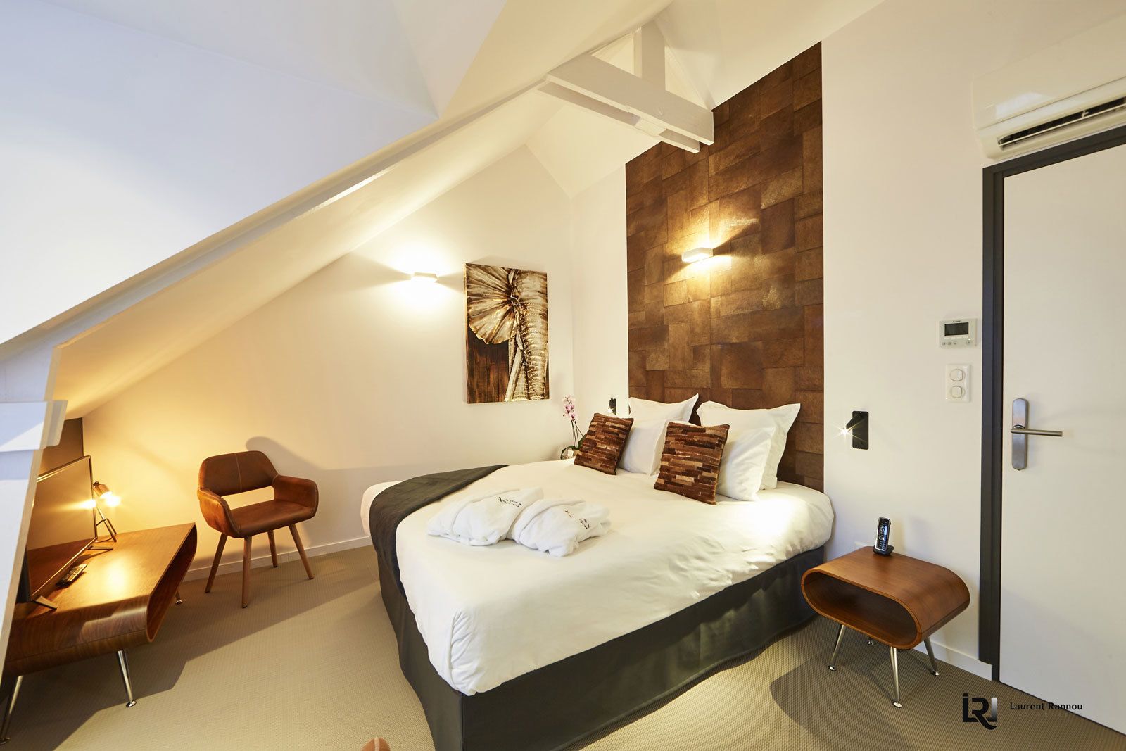 Ein modernes Schlafzimmer mit schrägen Wänden, einem Bett mit weißer Bettwäsche, einem Holz-Kopfende und einem Schreibtisch mit Stuhl.