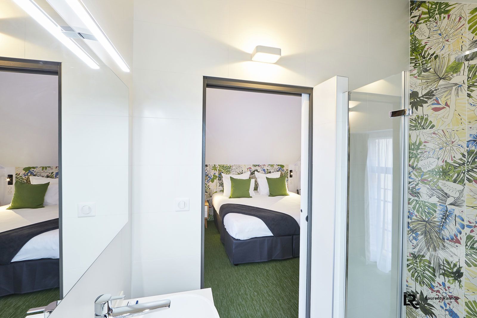 Una habitación de hotel con un espejo grande, una cama con ropa de cama verde y blanca, una mesita de noche con una lámpara, y un baño con un lavamanos y espejo.