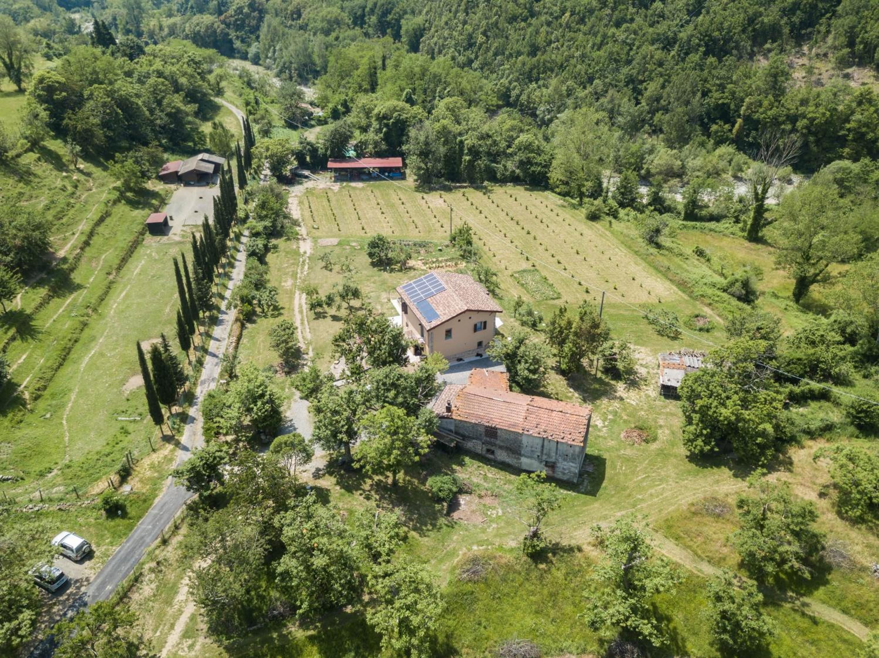 Vue aérienne d’une propriété rurale comprenant plusieurs bâtiments, dont une maison équipée d’un panneau solaire, le tout entouré de champs et d’arbres.