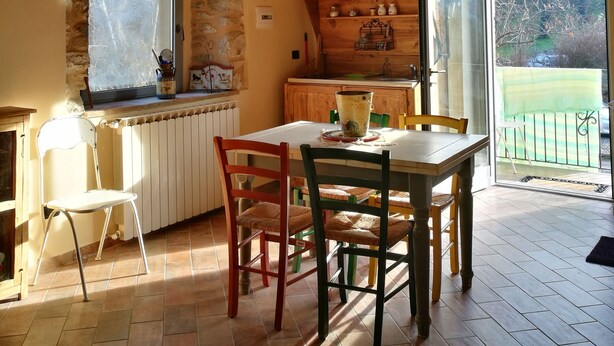 A dining area with a chandelier, stone wall, wooden ceiling, and a view of a green landscape through a sliding door. - Podere Groppini (Fivizzano)