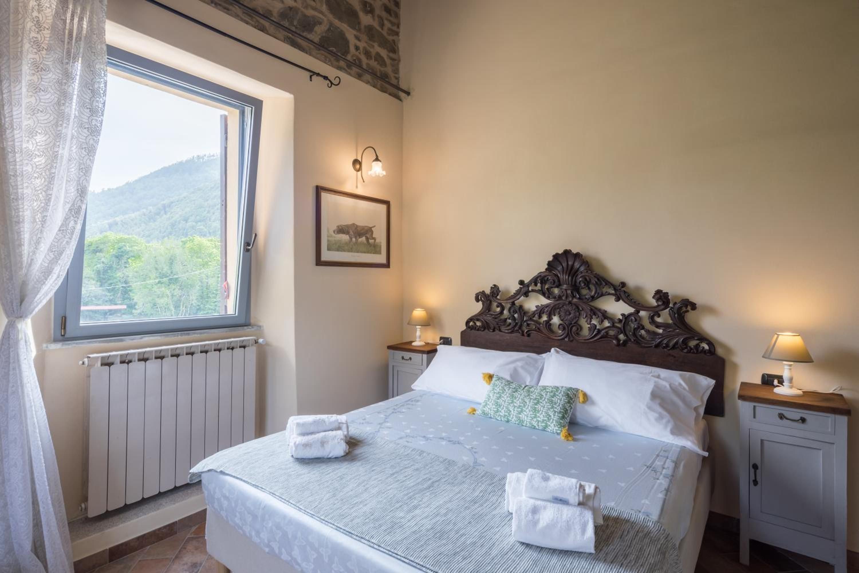 Una cama bien tendida con sábanas blancas y dos almohadas, un cuadro en la pared y una ventana con vistas a zonas verdes.