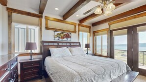 Een slaapkamer met een groot bed, houten hoofdbord, twee nachtkastjes met lampen, een bankje en grote ramen met zonwering.
