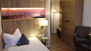Quarto de hotel com uma cama, mesa de cabeceira com abajur, guarda-roupa de madeira e uma cadeira.