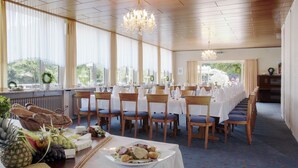 Uma sala de jantar com mesas grandes dispostas para uma refeição, cadeiras e um buffet com comida e bebidas.