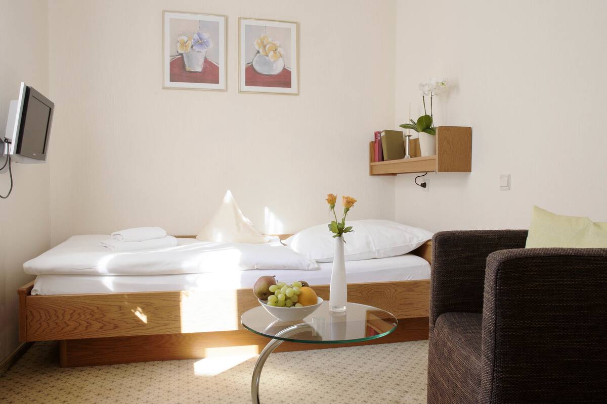 A neatly arranged bedroom with a bed, bedside table with fruit, a vase, and a chair.