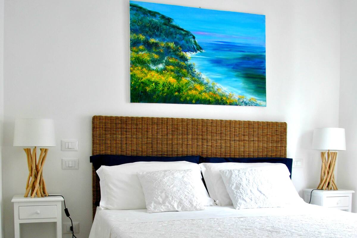 A bedroom with a bed, white nightstands, a wicker headboard, and a painting of a coastal landscape.