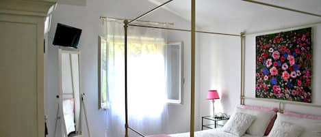 Quarto com cama de dossel, pintura de flores na parede, abajur e janela com cortinas.