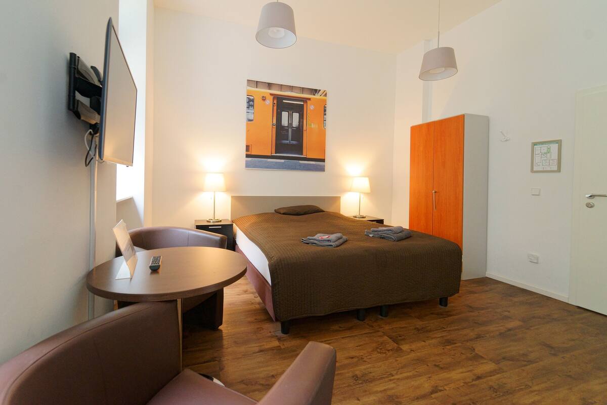 Una habitación de hotel con cama, escritorio, silla, armario y televisor instalado en la pared.