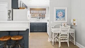 A kitchen with dark cabinets, a white countertop, and a window with a view of buildings. There is a dining area with white wicker furniture and a painting on the wall.