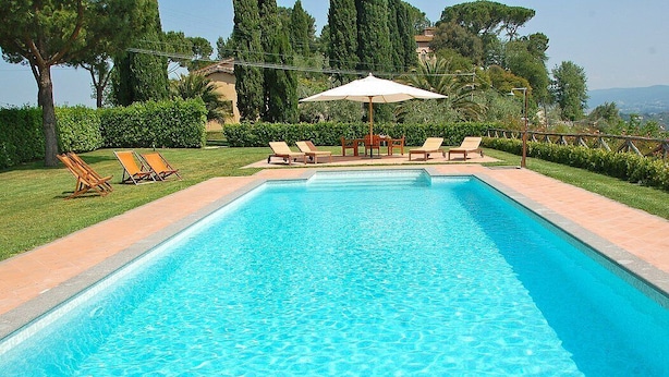 Une piscine rectangulaire à l’eau d’un bleu limpide, entourée d’une terrasse carrelée. On y trouve des chaises longues et une table avec un parasol.