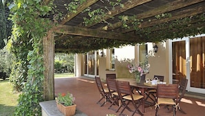 Une terrasse extérieure dotée d’une pergola en bois recouverte de lierre, une table ronde avec des chaises et un vase rempli de fleurs.