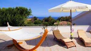 A hammock, two lounge chairs, and an umbrella on a terrace with a view of the sea.