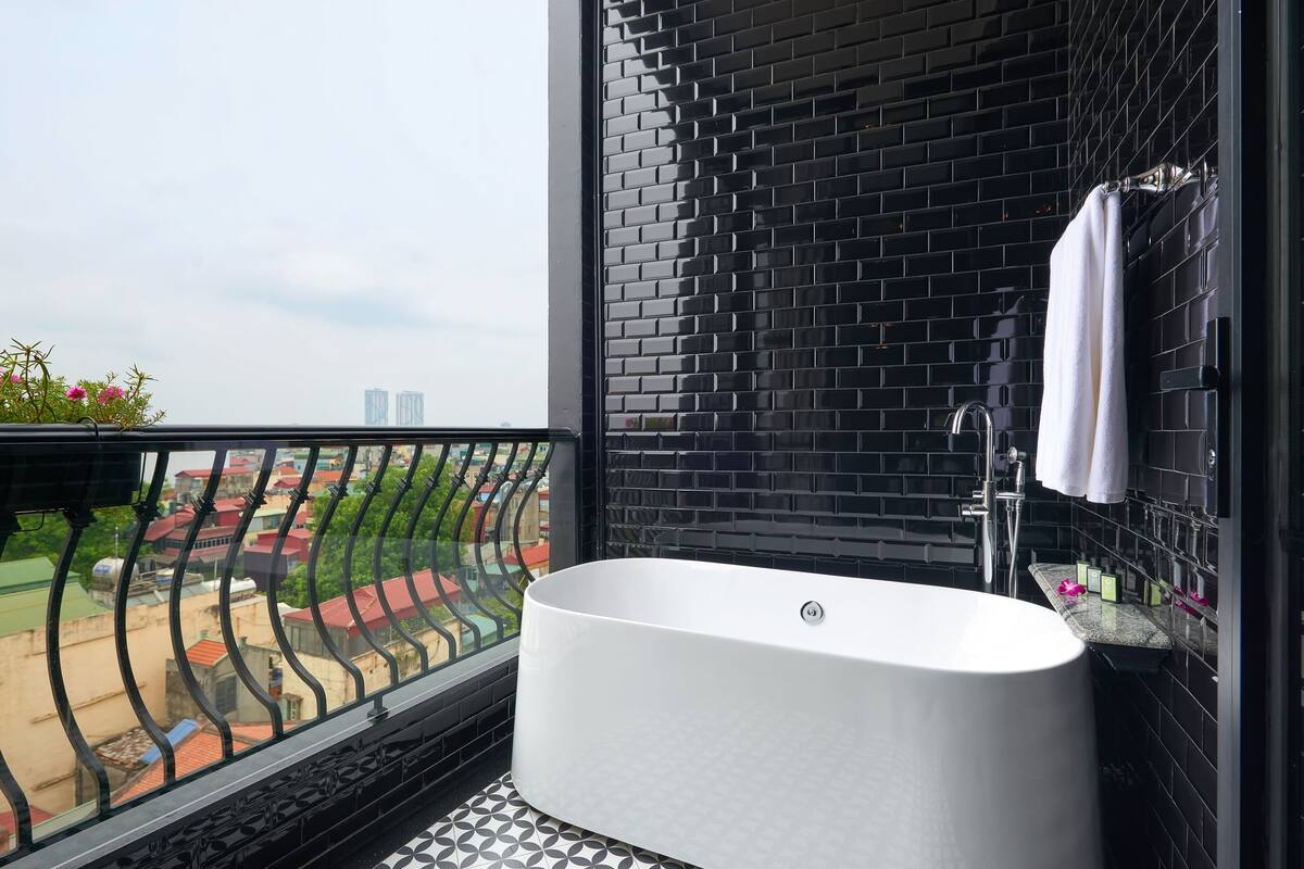 A modern bathroom with a freestanding bathtub, black tiled walls, and a balcony with a view of buildings.