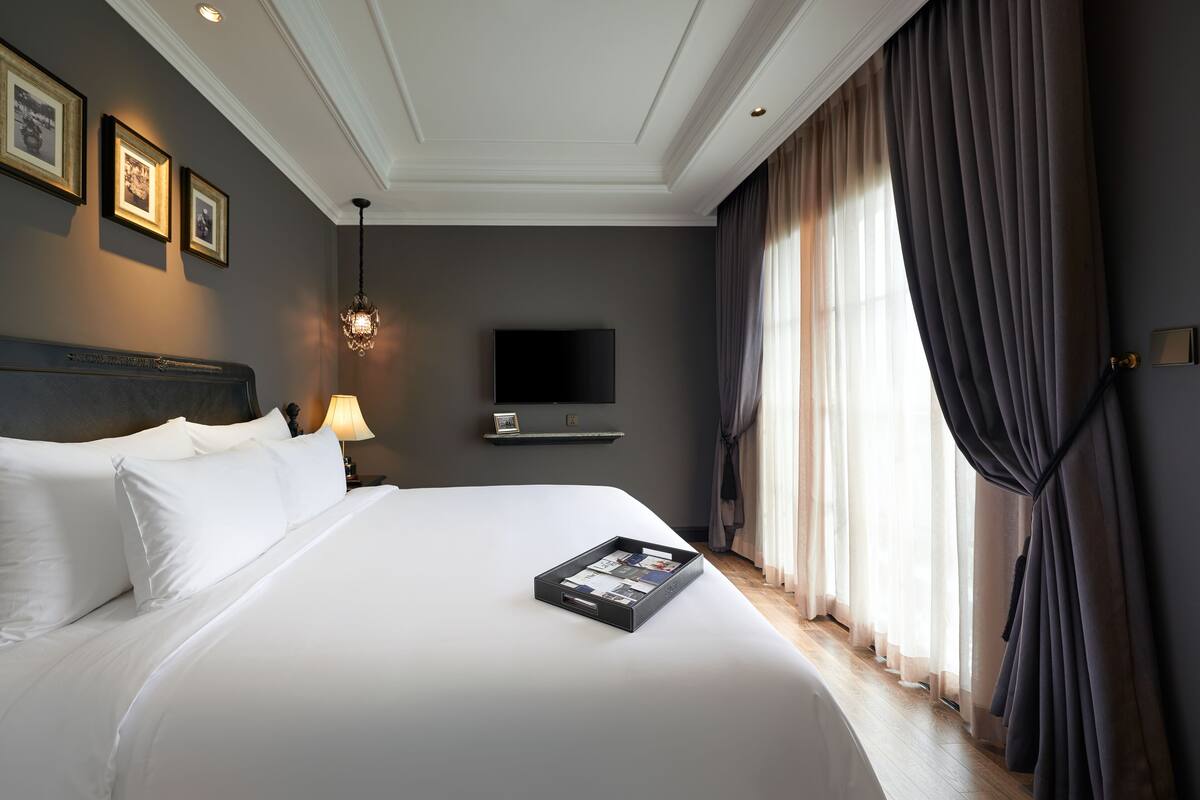 A neatly made bed with white linens, a tray of magazines, a television on the wall, and a large window with curtains.