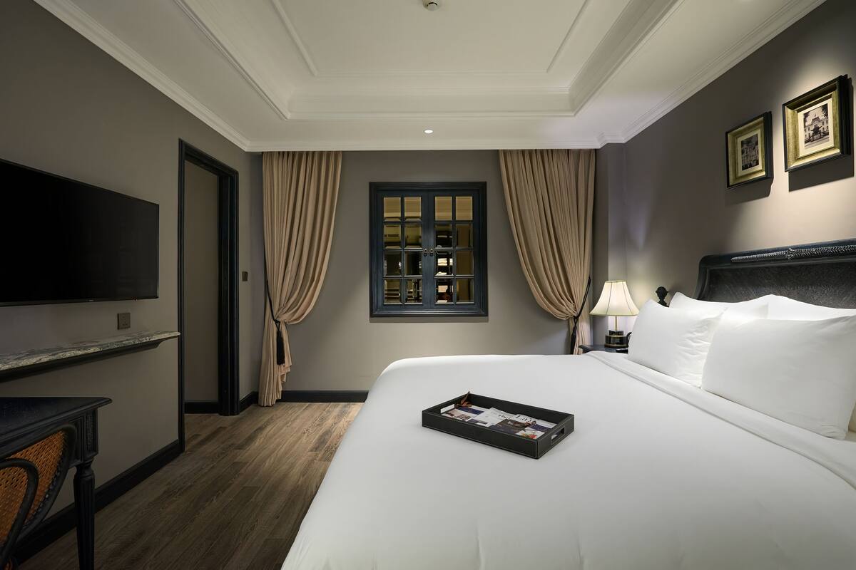 A hotel room with a large bed, a flat-screen TV mounted on the wall, a desk with a chair, and a window with curtains.