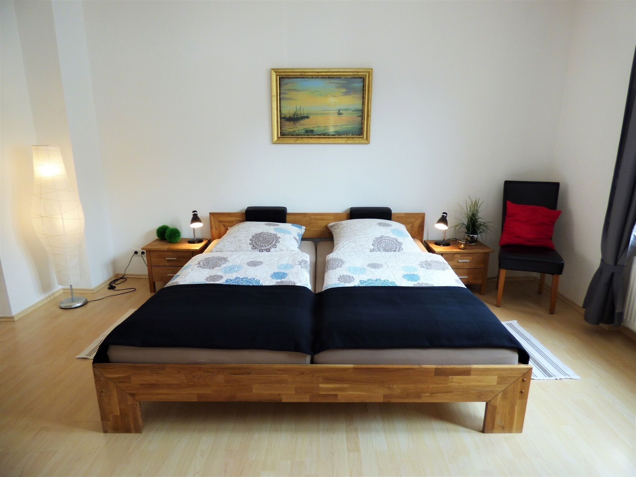 A double bed with a wooden frame, two bedside tables with lamps, a chair with a red cushion, and a painting on the wall.
