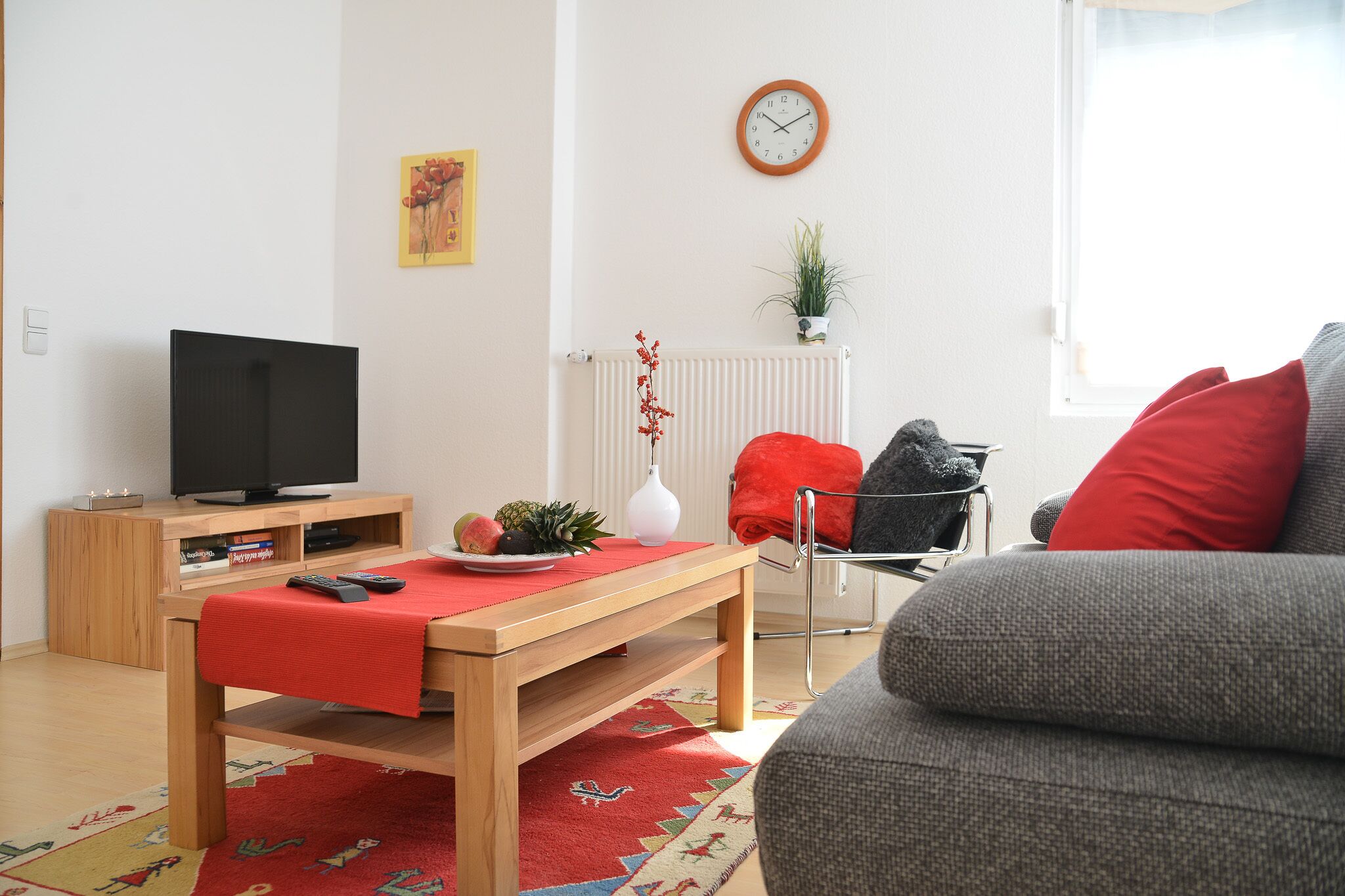 Una sala de estar moderna con un mueble bajo de televisión de madera, una mesa roja y un sofá gris.