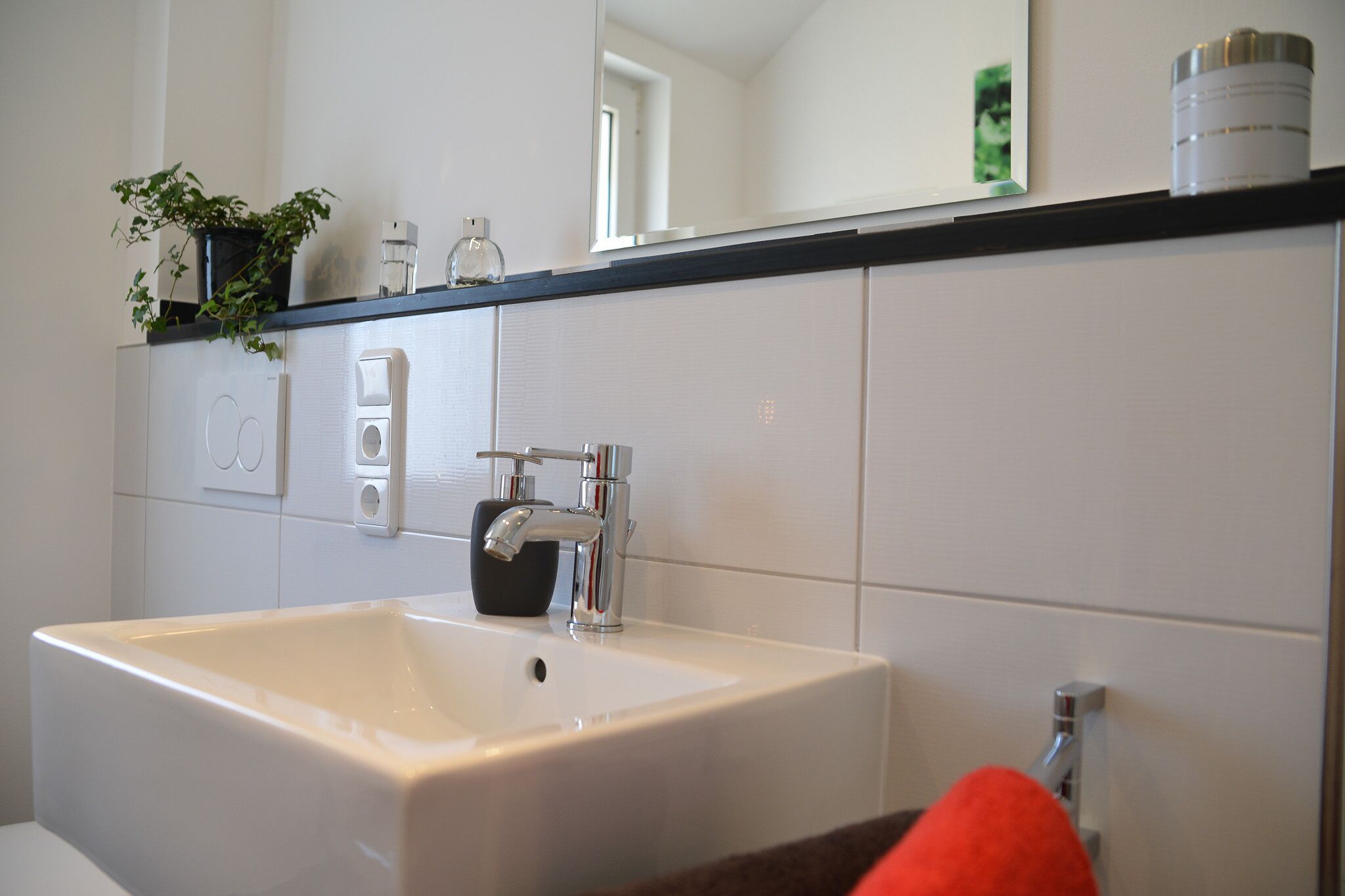 Een badkamer met een witte wastafel, een spiegel en een zwart aanrechtblad met een plant en een zeepdispenser.