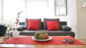 A living room with a grey sofa, red cushions, a white vase with red berries, a plate with fruits, and two remote controls on a red placemat. - Sonnenhaus Ferienwohnungen (Traben-Trarbach)