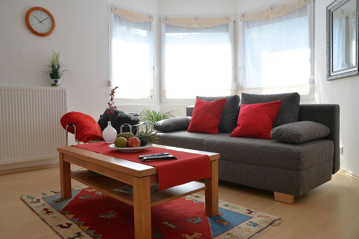 Ett vardagsrum med en grå soffa, röda kuddar, ett träbord och en klocka på väggen.