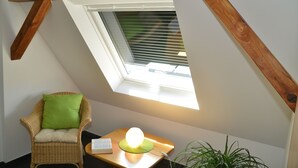 A small, cozy room with a sloped ceiling, a window with blinds, a wicker chair with a green cushion, a small table with a lamp, and a potted plant. - Sonnenhaus Ferienwohnungen (Traben-Trarbach)