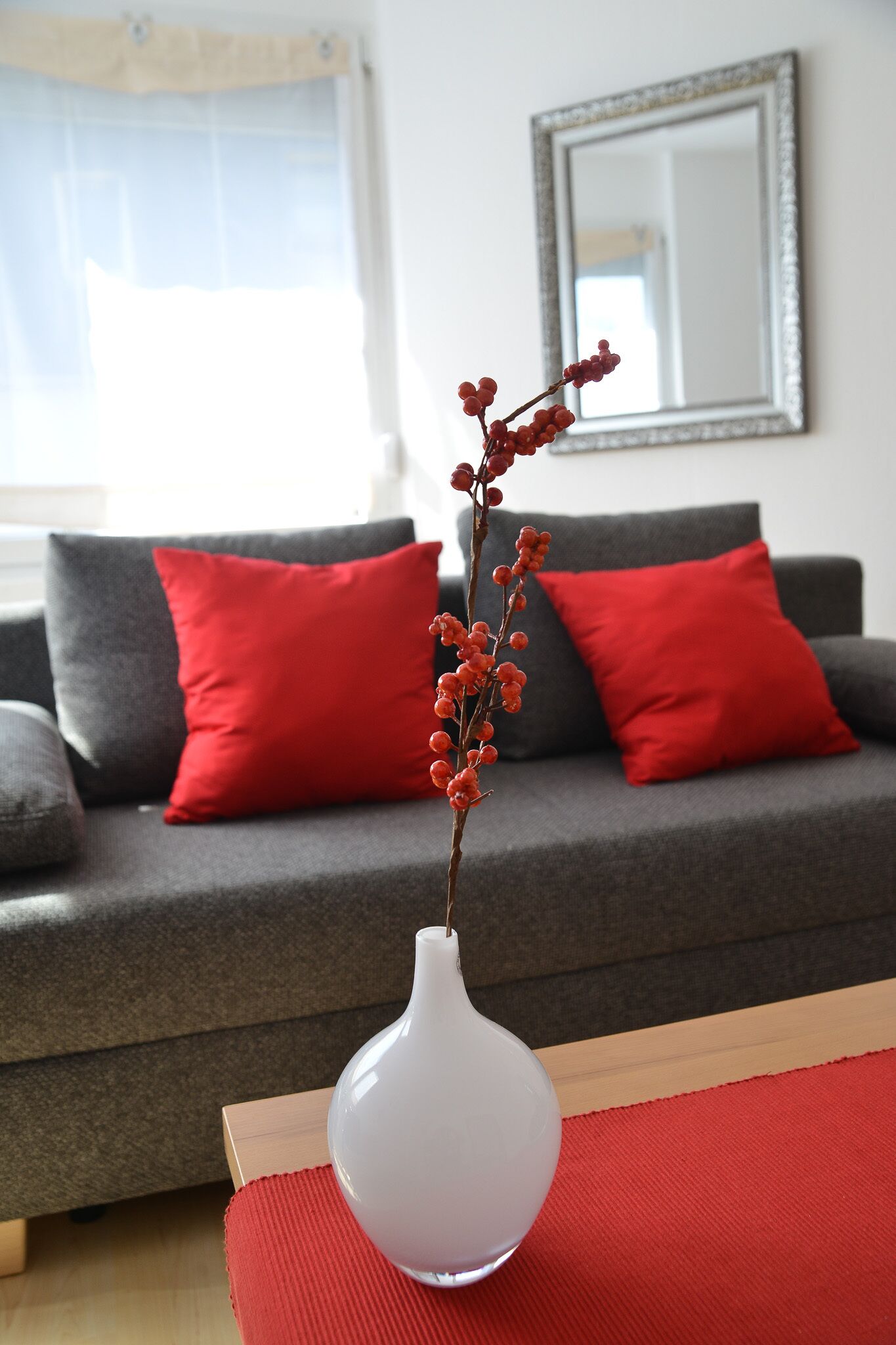 Un salon avec un canapé gris, des coussins rouges et un vase blanc contenant une branche, le tout posé sur un tapis rouge.