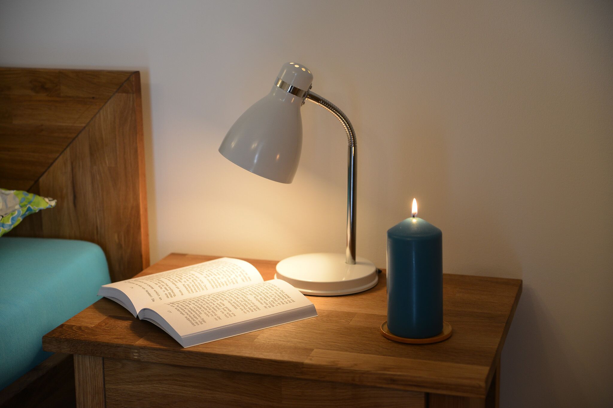A wooden bedside table with a white desk lamp, a lit blue candle, and an open book.