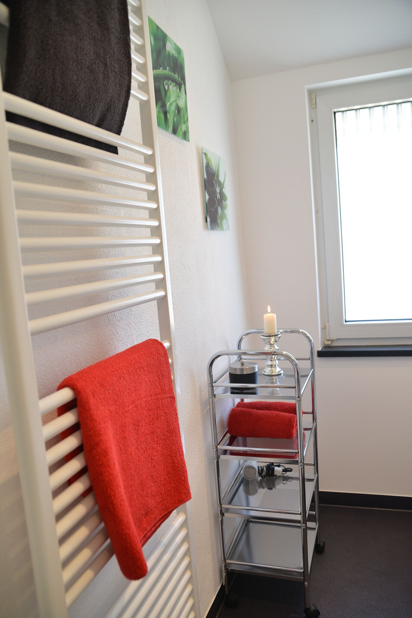 Une pièce avec un radiateur blanc, une serviette rouge accrochée dessus, un chariot en métal avec une bougie, et une fenêtre avec des stores.
