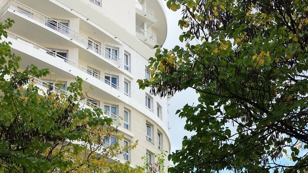 Un edificio de varios pisos con balcones, rodeado de árboles con hojas verdes y amarillas.