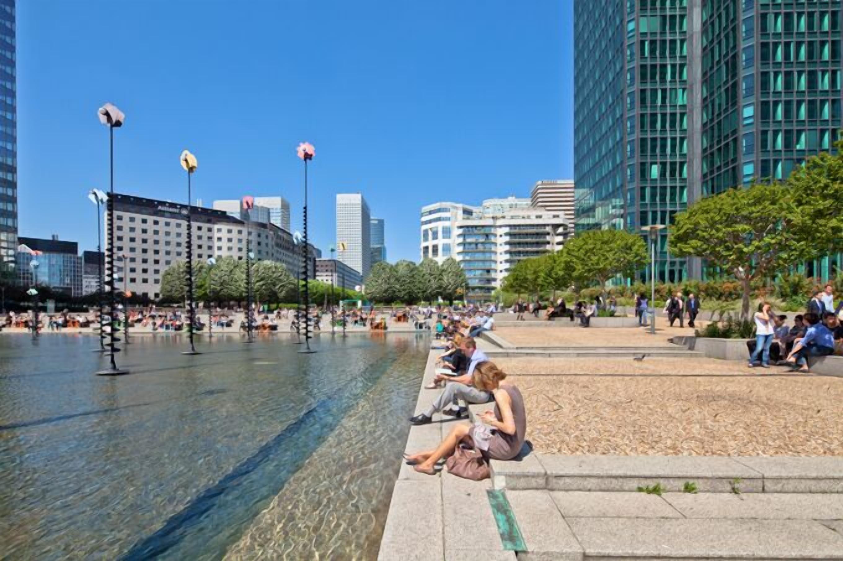 Une place publique agrémentée d’une fontaine, de coins salon et bordée de bâtiments modernes.