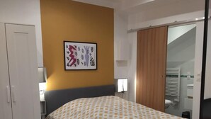 Une chambre à coucher comprenant un lit, un bureau, une chaise, une armoire et une salle de bain visible à travers une porte coulissante.