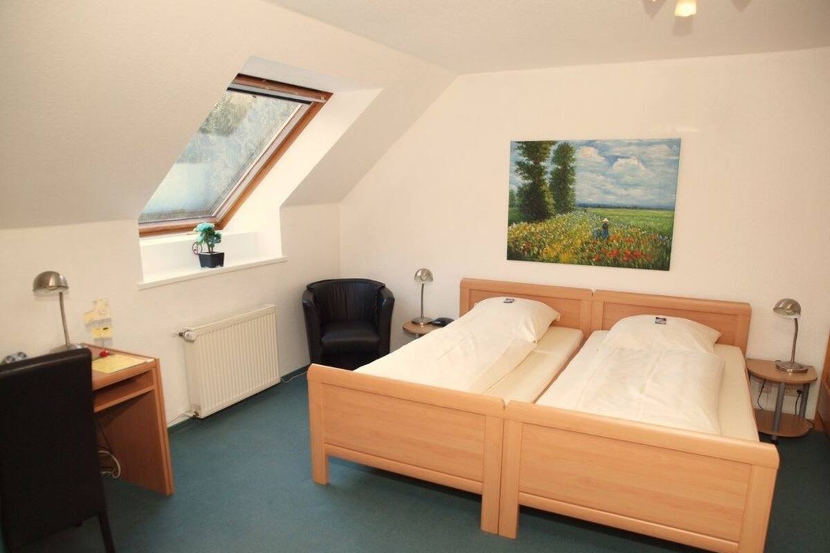 Un dormitorio con una cama de madera, un escritorio, una silla, un radiador, una ventana con una planta y un cuadro en la pared.