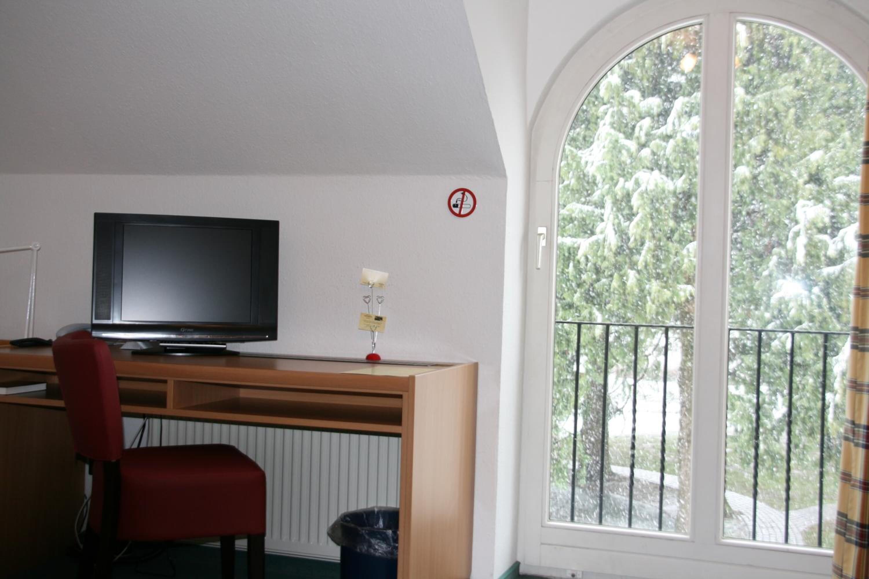 A room with a TV on a wooden desk, a red chair, a window with a view of trees, and a no smoking sign.