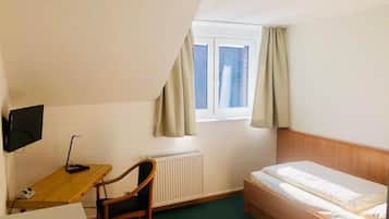A small, single-room hotel room with a bed, a desk, a chair, a radiator, and a window with curtains.
