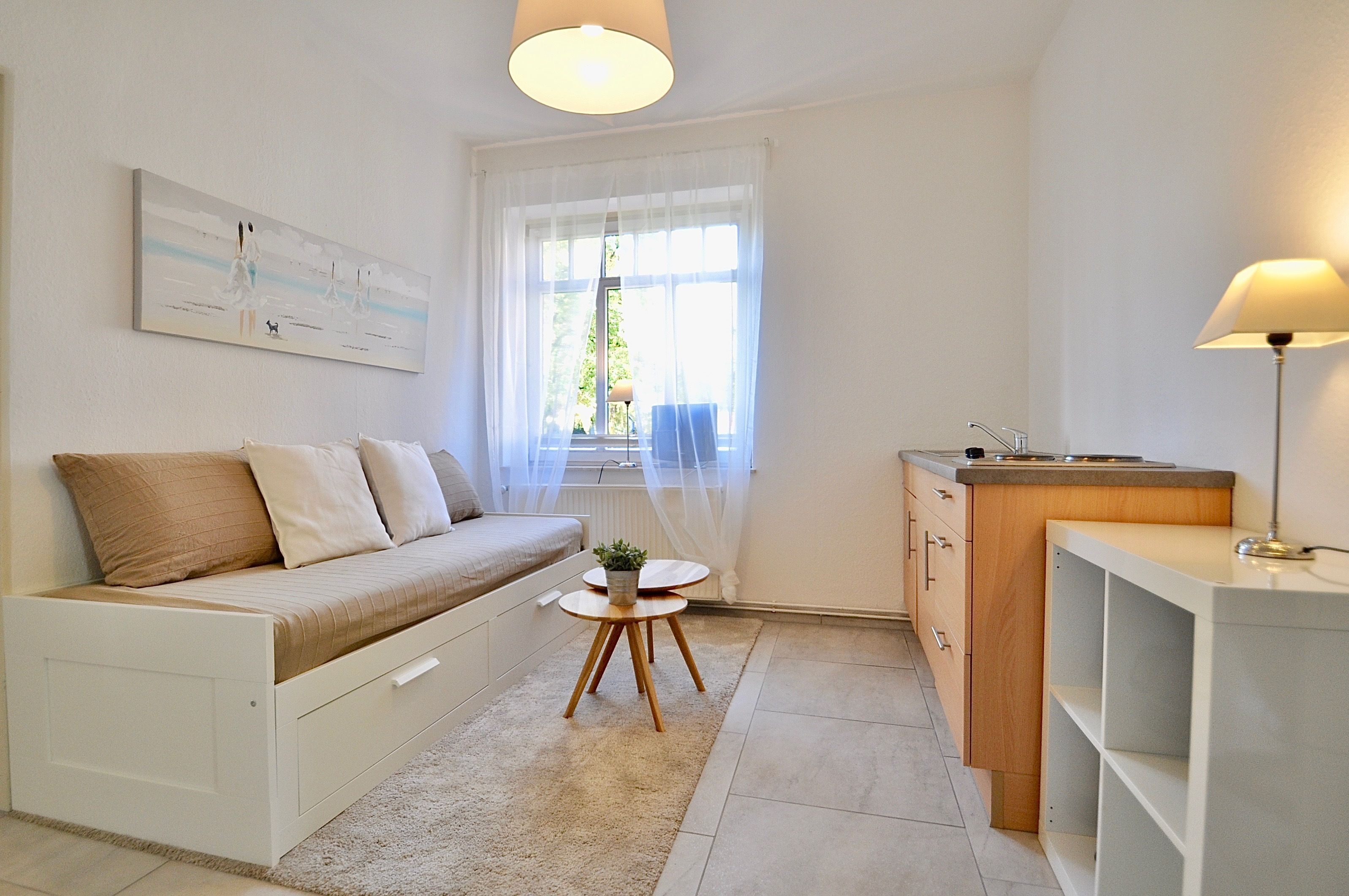 Un salon moderne et minimaliste, comprenant un canapé, une petite table ronde avec une plante, un coin cuisine avec un évier et des placards, et une grande fenêtre ornée de rideaux légers.